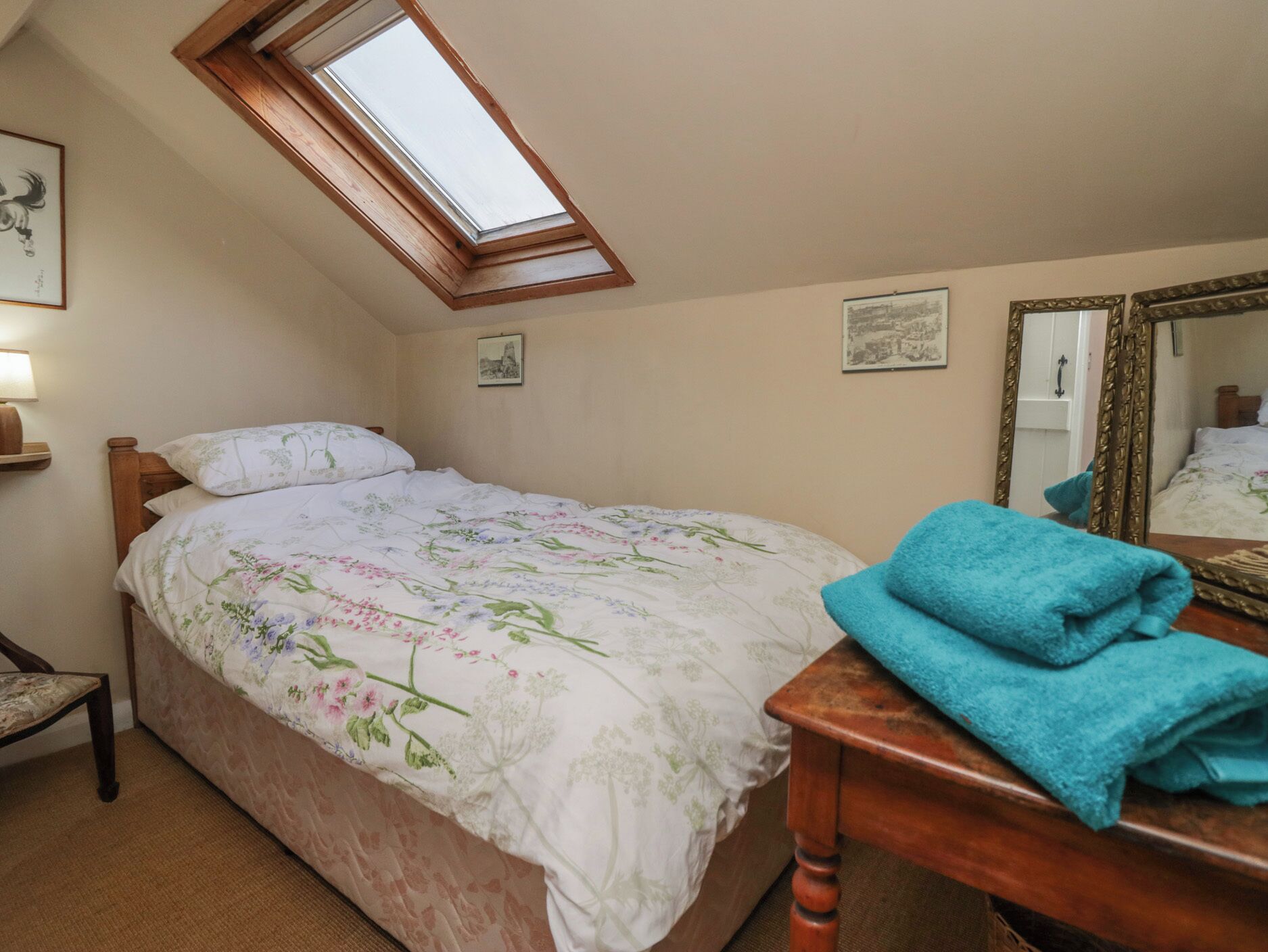 A bedroom with a sloped ceiling, a bed with floral bedding, a wooden nightstand, a blue towel, and a mirror with an ornate frame.