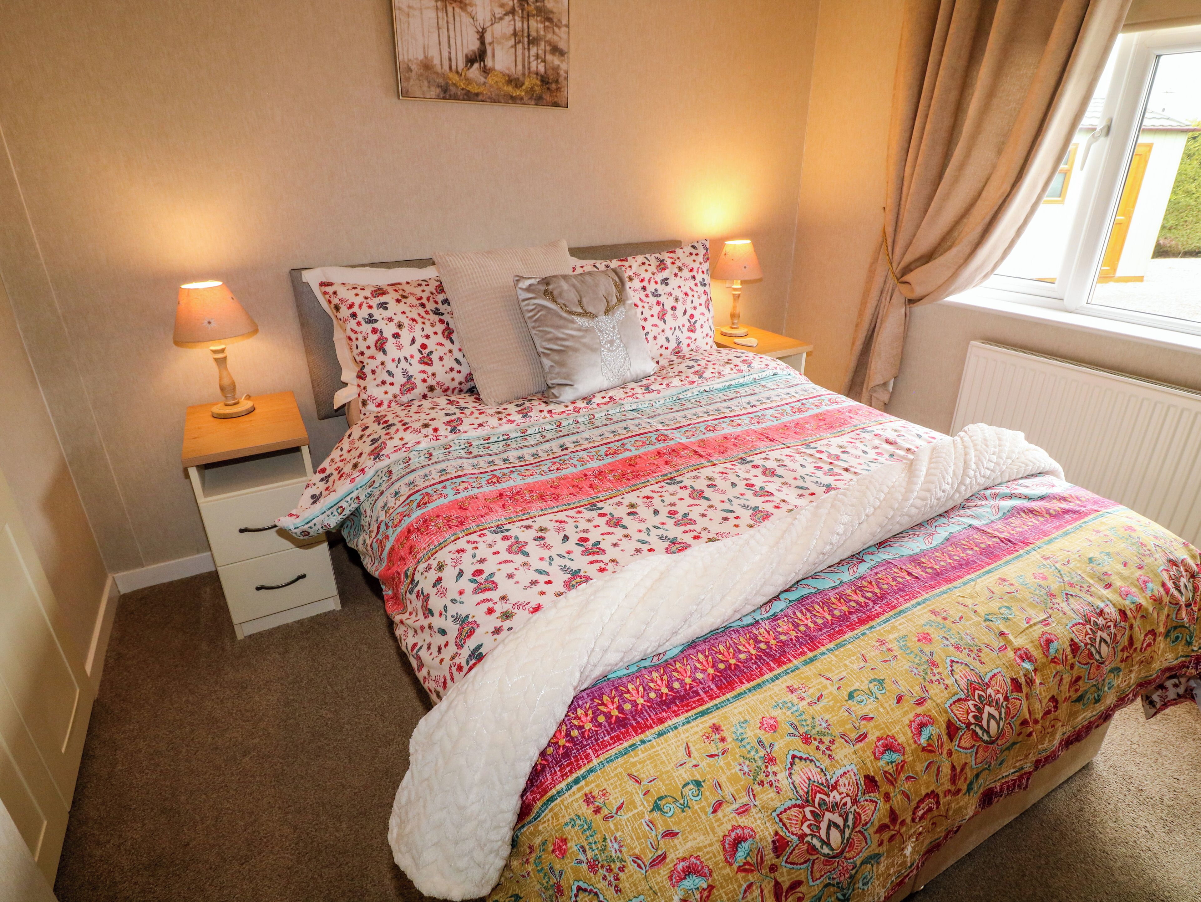 A bedroom with a bed, two bedside tables, a lamp on each table, a window with curtains, and a wall-mounted painting.