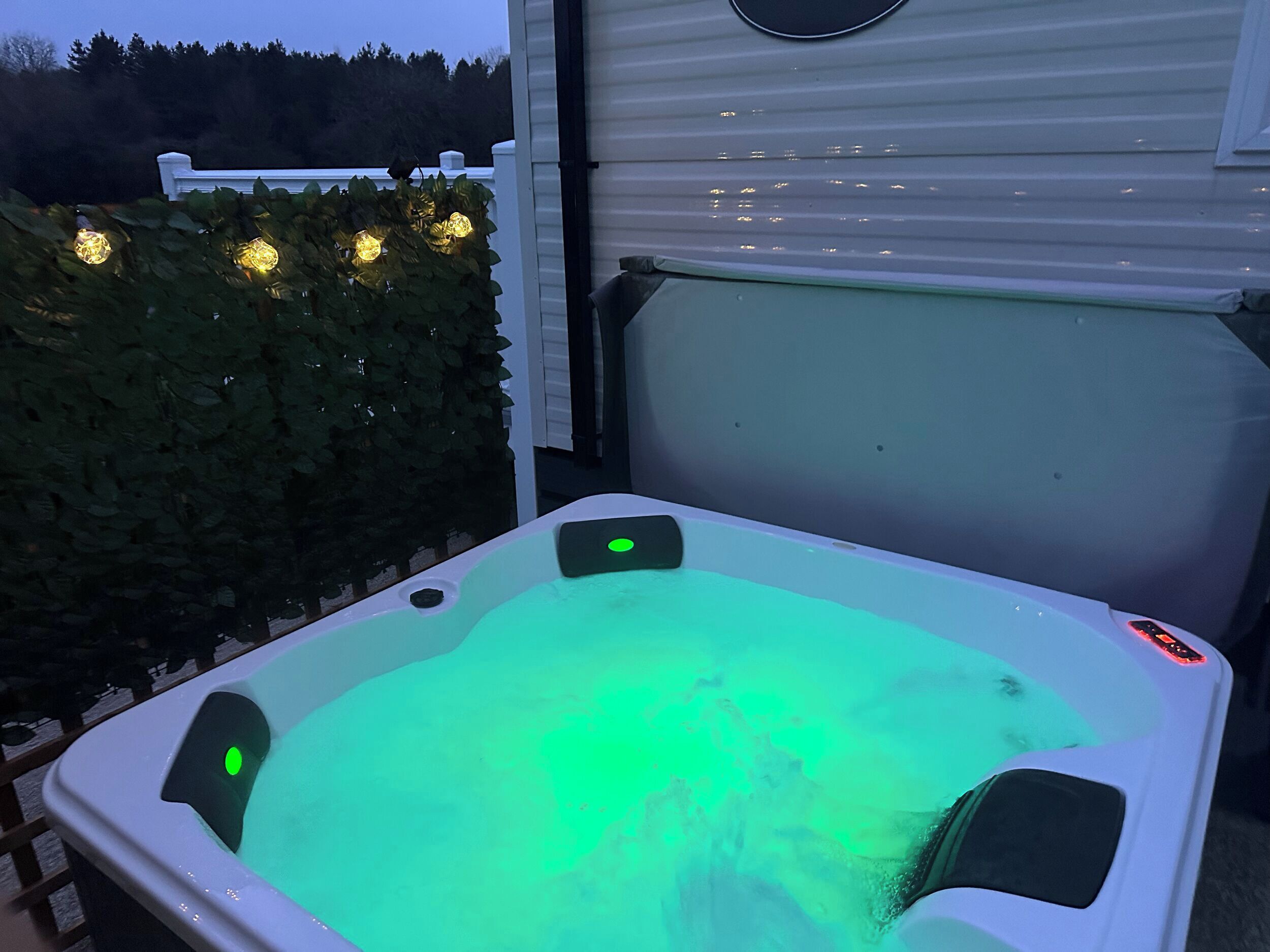 A hot tub with a green light, surrounded by a white fence and a hedge.