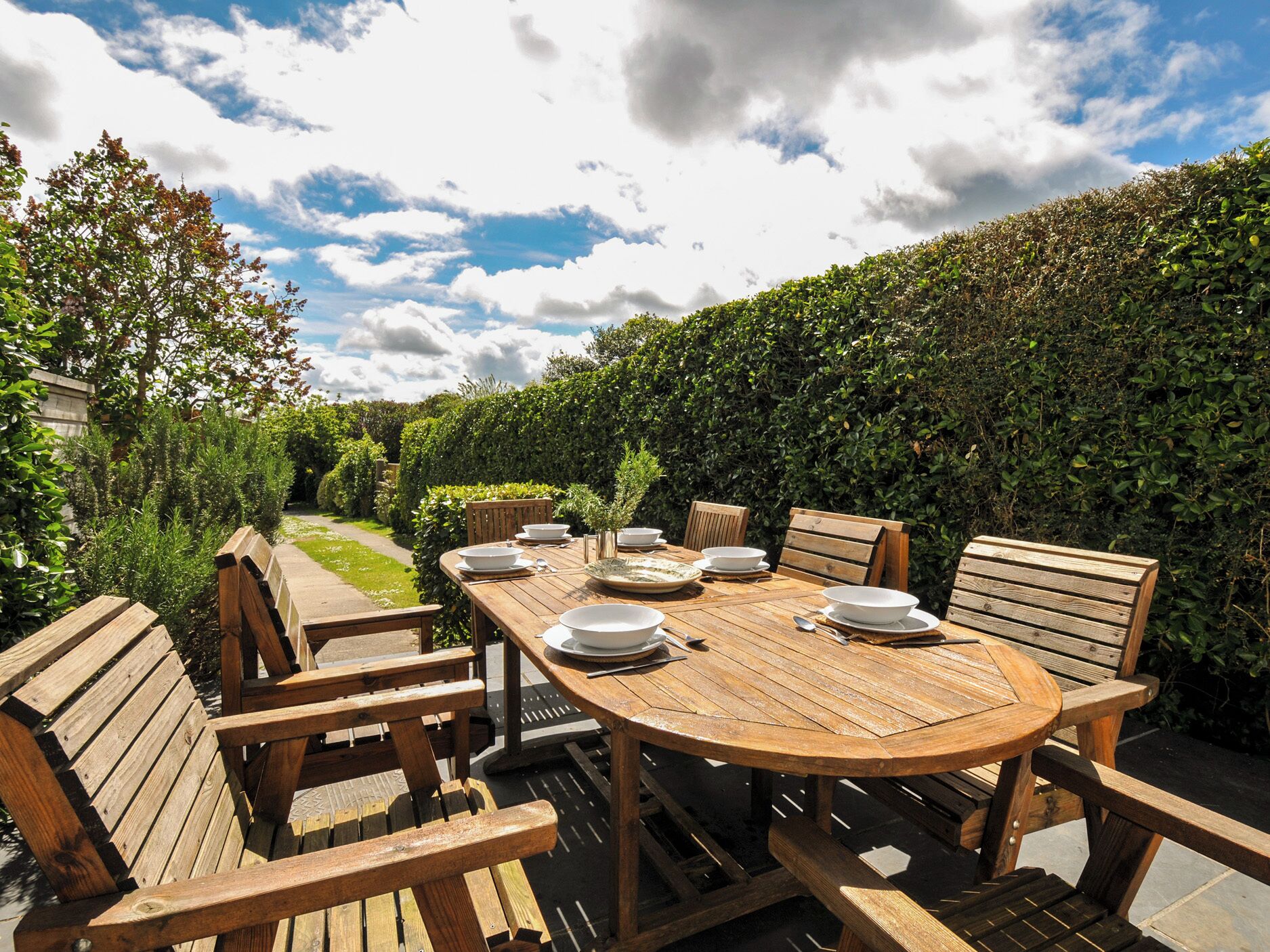 A wooden outdoor dining table with chairs, surrounded by a hedge and a garden path.