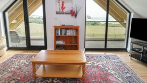 Une pièce spacieuse avec une grande table en bois, une bibliothèque et un téléviseur. Elle est dotée d’une grande baie vitrée donnant sur l’extérieur.
