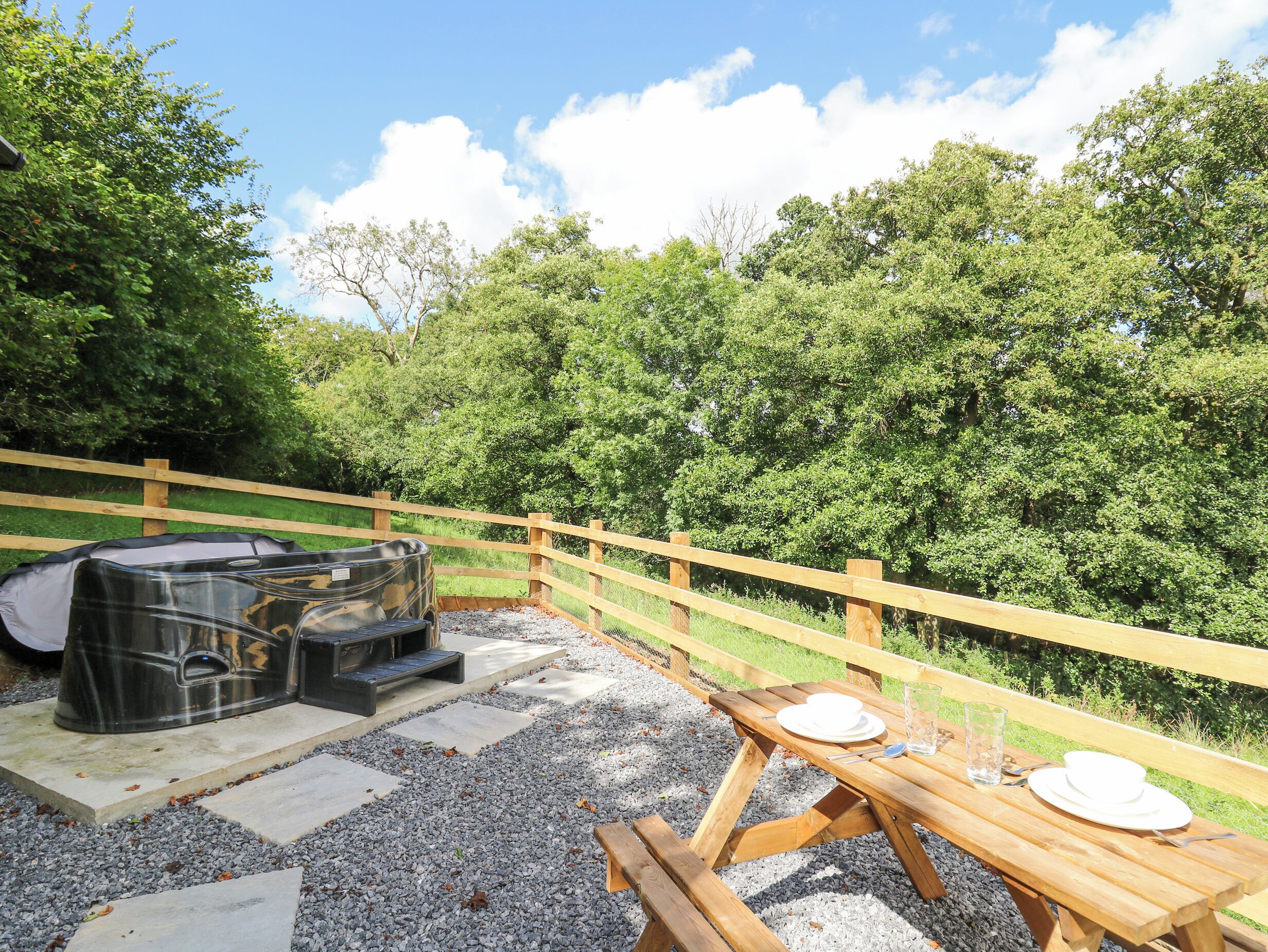 An outdoor patio with a hot tub, picnic table, and seating area surrounded by a wooden fence and lush greenery.