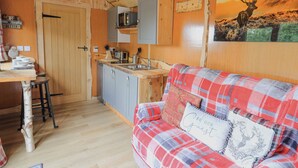 Interior acogedor de una cabaña con una mesa de comedor de madera, una kitchenette y un sofá escocés con un cojín que dice 'BE OUR Guest'.