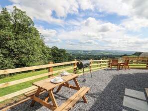 A wooden picnic table with a grill and chairs, surrounded by a wooden fence and gravel, overlooking a scenic landscape. - Shepherd's Cwtch (Llandovery)