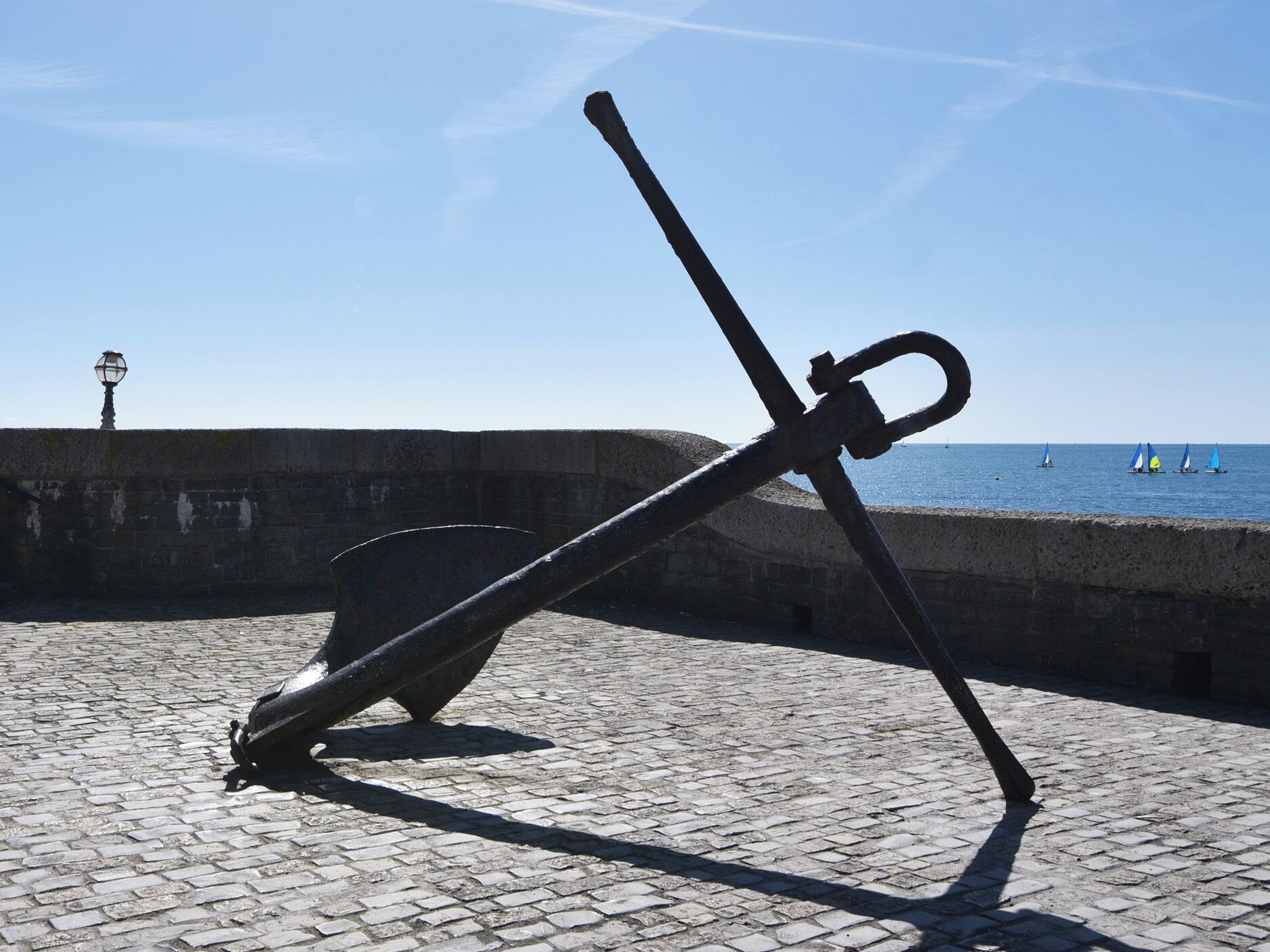 Une grande sculpture d’ancre est installée sur un pavé de pierres, au bord de l’eau, près d’un mur de pierres et d’un lampadaire.