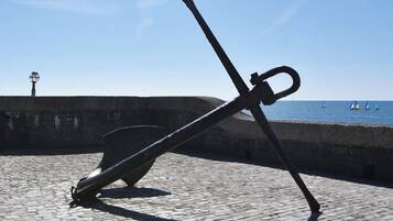 Une grande sculpture d’ancre est installée sur un pavé de pierres, au bord de l’eau, près d’un mur de pierres et d’un lampadaire.