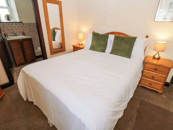 Una cama bien hecha con ropa blanca y almohadas verdes, una mesita de noche de madera con una lámpara, un aparador de madera y un espejo en la pared.