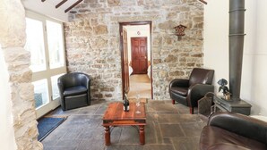 A stone-walled living room with two leather chairs, a wooden coffee table with a bottle and glass, and a fireplace. - Pencroft Cottage (Leyburn)