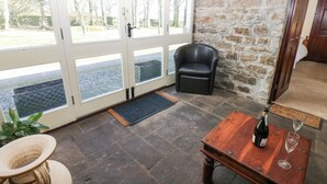 A cozy room with a stone wall, a black leather chair, a wooden table with a bottle and glass, and a large window with a view of trees. - Pencroft Cottage (Leyburn)