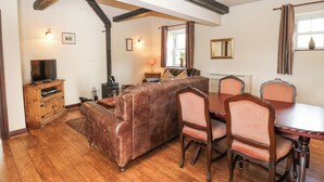 A cozy living room with a brown leather sofa, wooden dining table, and matching chairs. A television is on a wooden cabinet, and a fireplace is visible in the background. - Pencroft Cottage (Leyburn)