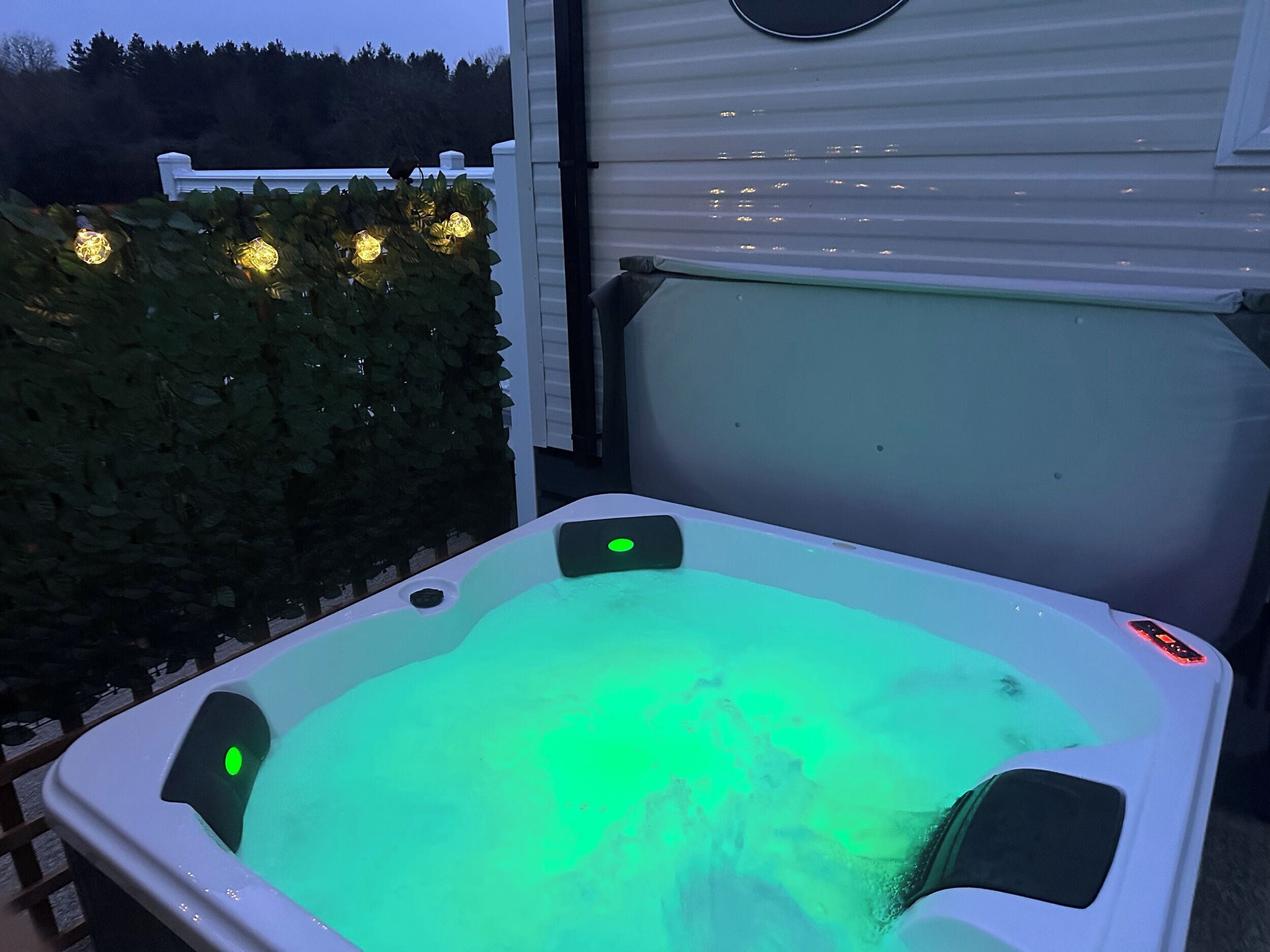 Ein Whirlpool mit grüner Beleuchtung, umgeben von einem weißen Zaun und einer Hecke.