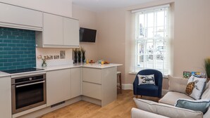Een moderne keuken met witte kasten, een blauwgroene spatwand en een ingebouwde oven. Er is een raam met jaloezieën, een blauwe stoel en een bank met decoratieve kussens.