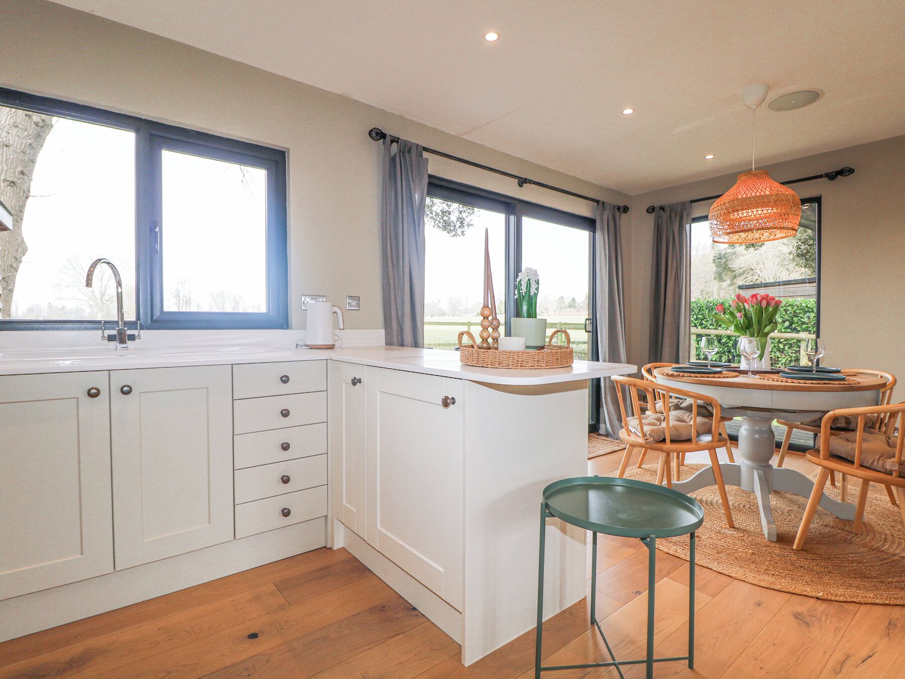 A modern kitchen with white cabinetry, a large window, and a dining area with wooden chairs and a table. - Holm Oaks Cottage (Godalming)