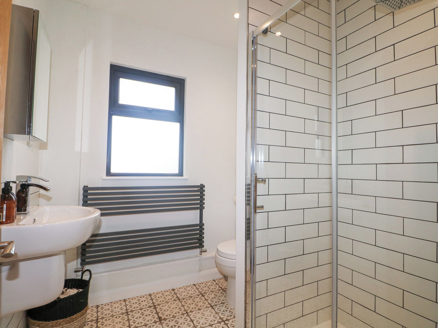 A bathroom with a white sink, a mirror, a window, a toilet, and a shower with white tiles.