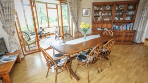 A dining area with a wooden table, chairs, and a vase of flowers. There is a bookshelf with books and decorative items. - Artist's Cottage (Penrhyndeudraeth)