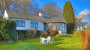 A house with a curved glass conservatory, a lawn with a dining set, and a hedge in the foreground. - Artist's Cottage (Penrhyndeudraeth)
