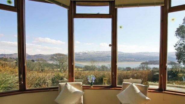 A room with a large window offering a view of a lake and mountains. There are two chairs with cushions. - Artist's Cottage (Penrhyndeudraeth)