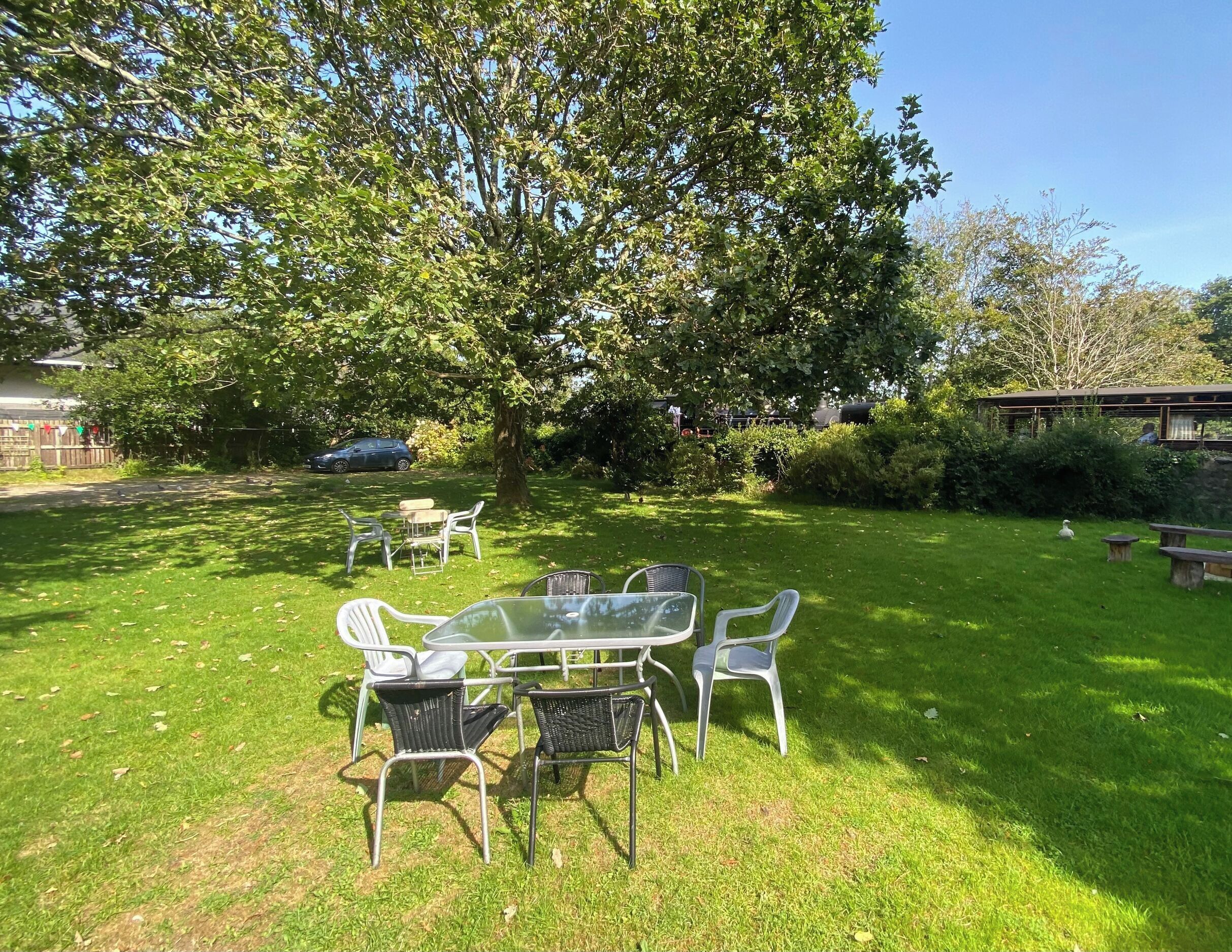 A grassy outdoor area with a table and chairs, surrounded by trees and a wooden structure.