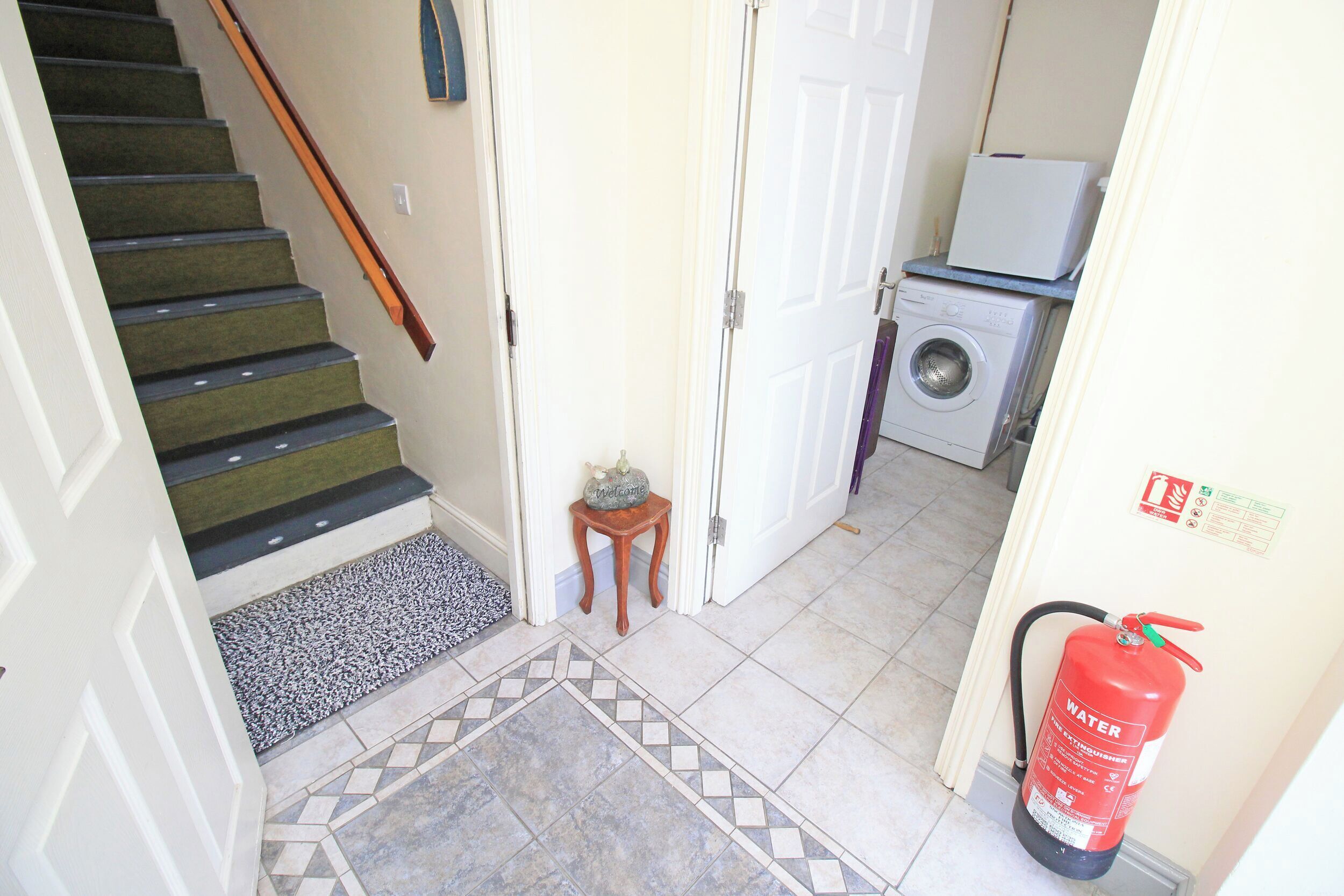 A hallway with a carpeted staircase, a small wooden stool, a washing machine, and a fire extinguisher.