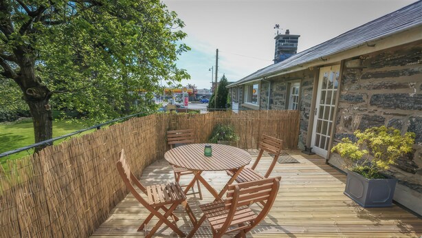 A wooden deck with a table and chairs, surrounded by a bamboo fence, overlooking a street with buildings and a clear sky. - Treetops Flat (Porthmadog)