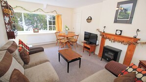 A living room with a sofa, a coffee table, a TV, a fireplace, a dining table, and a window with curtains. - Treetops Flat (Porthmadog)