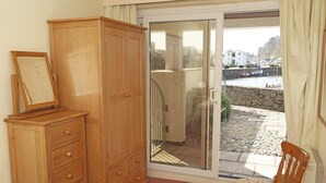 A room with a wooden wardrobe, a chair, and a sliding glass door leading to a balcony with a stone wall and a view of boats. - Glaslyn Harbour Views (Porthmadog)