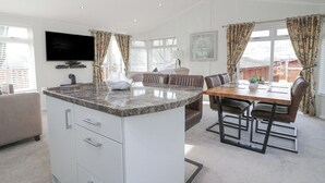 A modern kitchen with a granite countertop, white cabinetry, and a built-in island. There is a dining area with a wooden table and chairs, and a living room with a sofa and a flat-screen TV mounted on the wall. - Swifts Reach Lodge (Windermere)