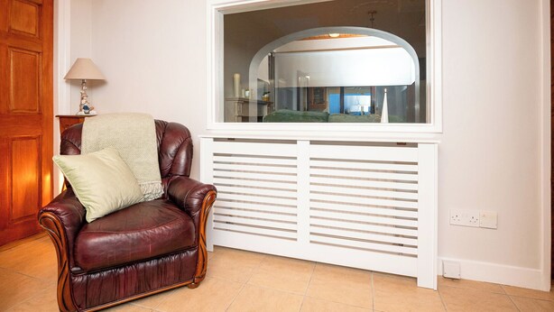 Une pièce avec un fauteuil en cuir marron, un radiateur blanc et une grande fenêtre donnant sur une cuisine.