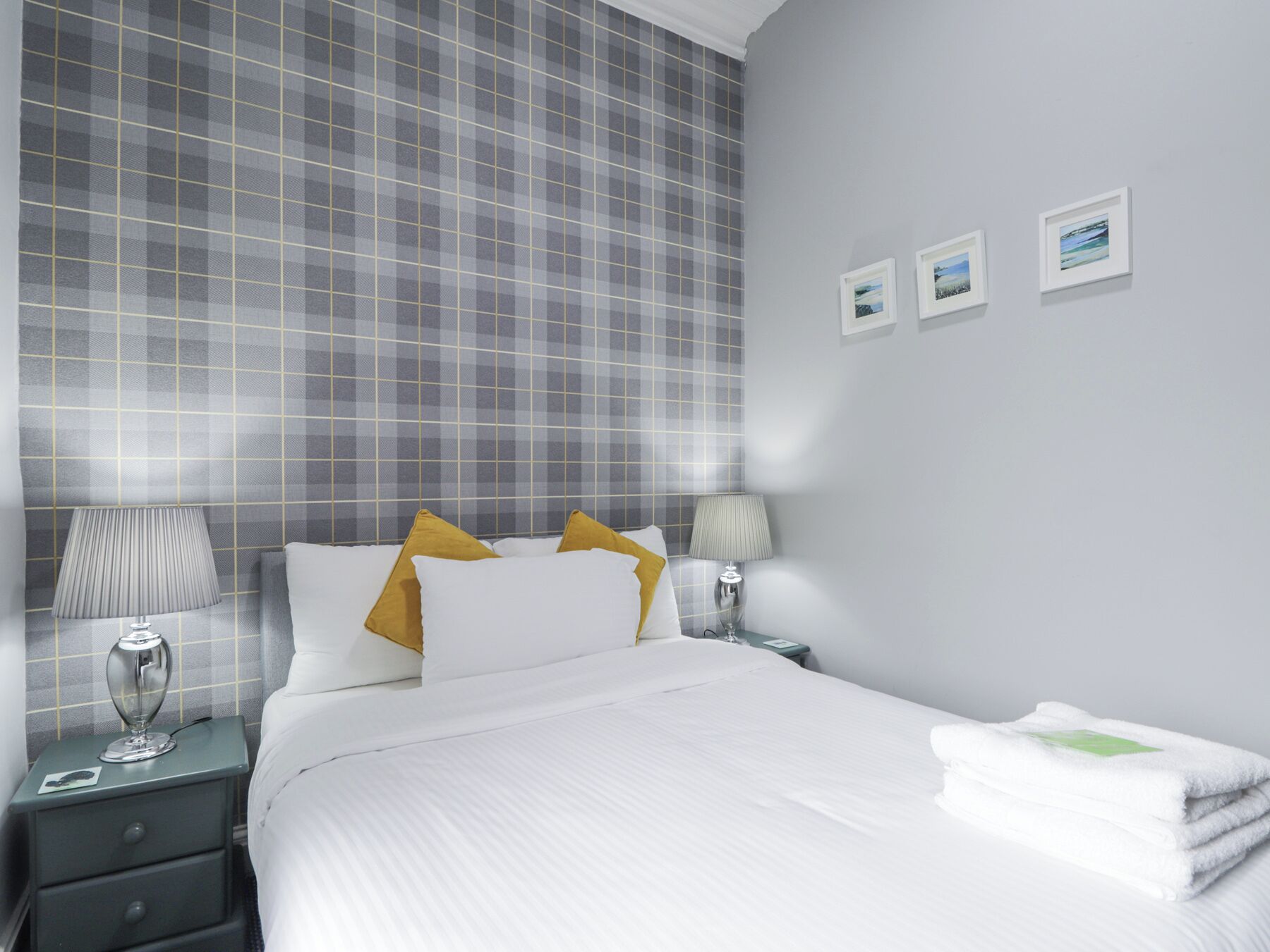 Een netjes ingerichte hotelkamer met een geruit behang, een bed met witte en gele kussens, een nachtkastje met een lamp en een aan de muur hangend fotolijstje.