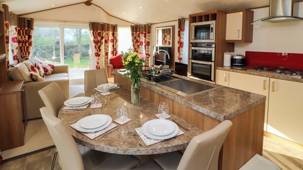 A modern kitchen with a central island, built-in oven, and microwave. There is a dining area with a table set for four, and a sofa in the background. - Lodge 4 (Carnforth)