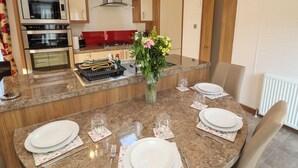 A modern kitchen with a central island, built-in oven, and a dining area set for four. - Lodge 4 (Carnforth)