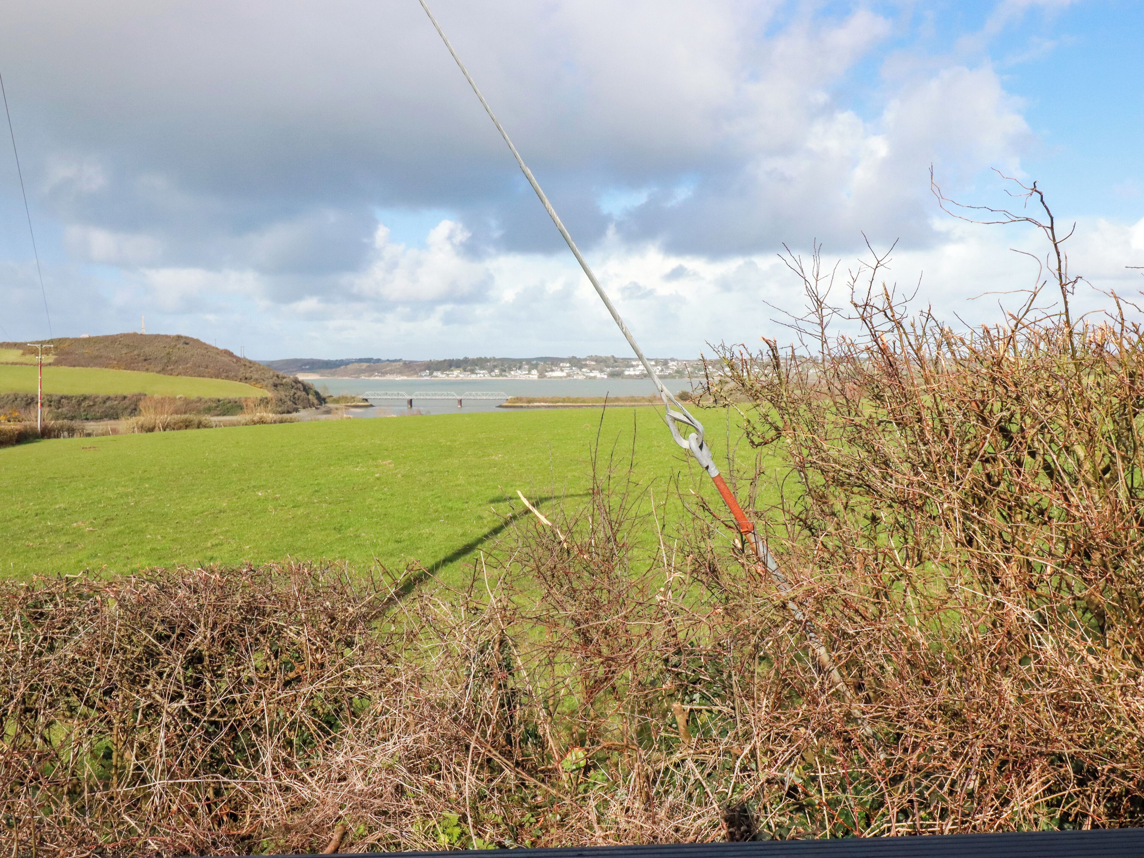A view of a green field with a bridge in the distance, a body of water, and a cloudy sky.