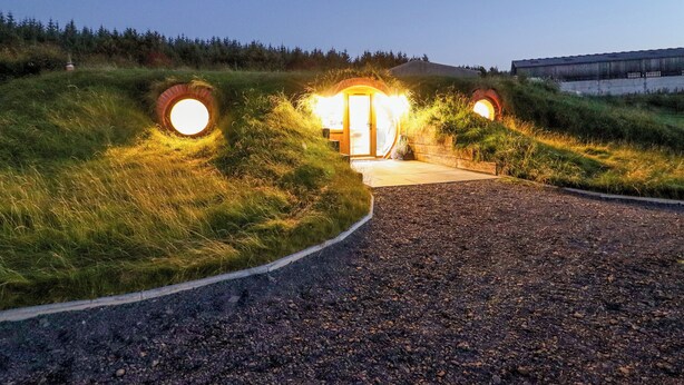 A pathway leading to a building with circular, illuminated entrances surrounded by grassy terrain. - Weirglodd (Ruthin)