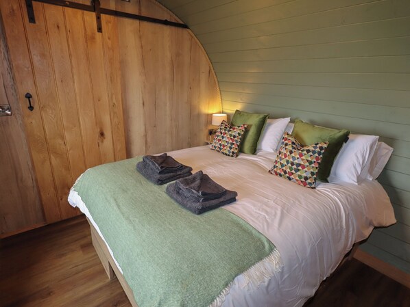 A bedroom with a wooden sloped ceiling, a bed with white linens, green and multicolored pillows, and a green throw blanket. - Weirglodd (Ruthin)
