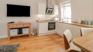 Una cocina moderna con armarios blancos, horno empotrado, microondas y cafetera. Hay una mesa de comedor de madera con sillas, un televisor montado en la pared y un taburete pequeño.