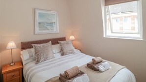 Un dormitorio ordenado con una cama, dos lámparas de noche, un cuadro enmarcado en la pared y una ventana con vistas a edificios.