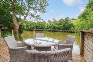 A wicker outdoor seating area with a glass table, overlooking a calm river and lush greenery. - 10 Water's Edge (Looe)