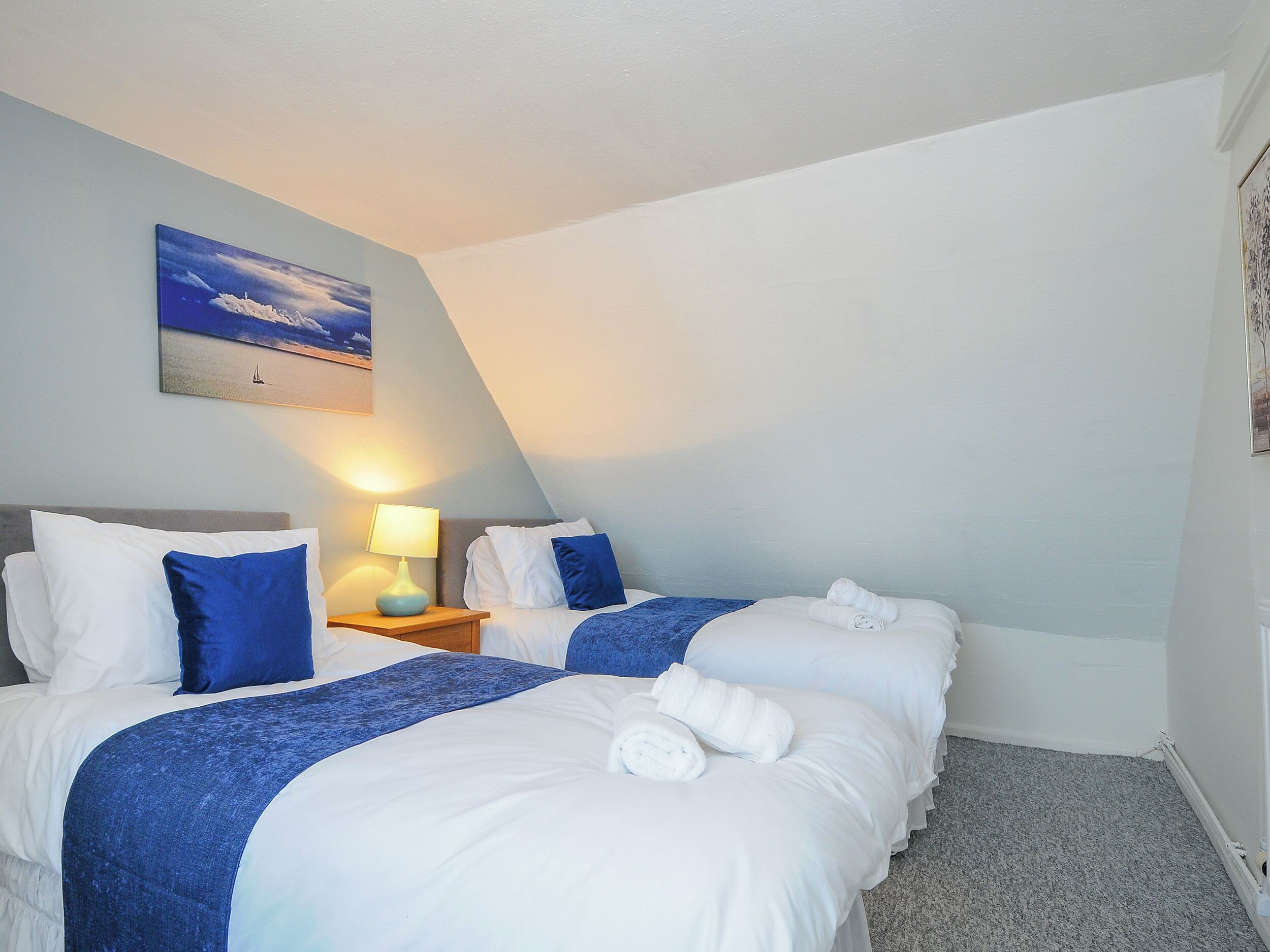 Habitación de hotel con dos camas, una mesita de noche con lámpara, un cuadro de un paisaje marino y piso alfombrado.