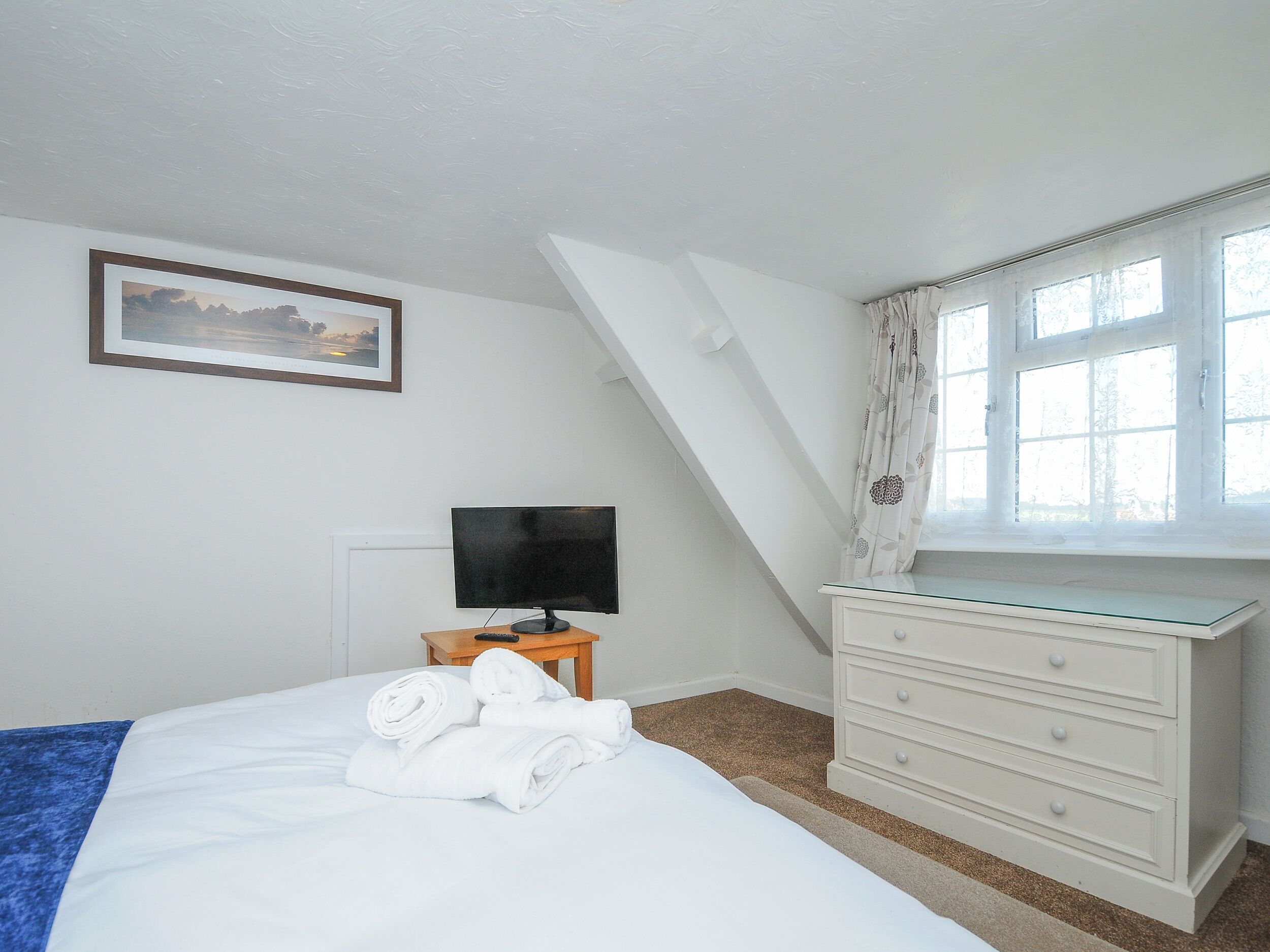 A bedroom with a bed, a TV on a wooden table, a dresser, and a window with curtains.