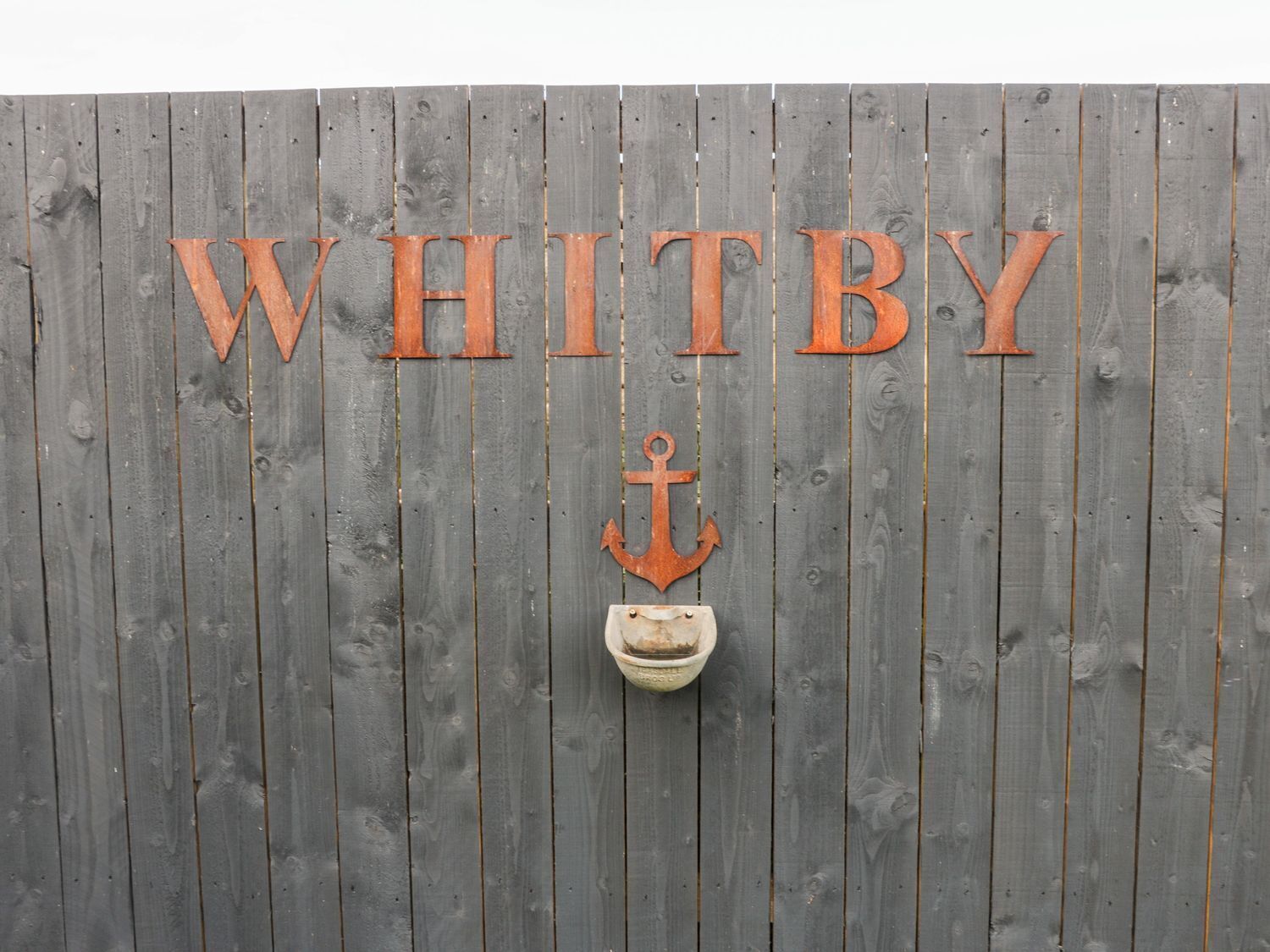 Een houten wand met de naam “WHITBY” in grote letters en een anker dat eronder hangt.
