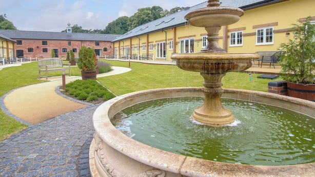 A courtyard with a stone fountain, a bench, and a building with multiple windows and doors. - Cheltenham Cottage (Oswestry)