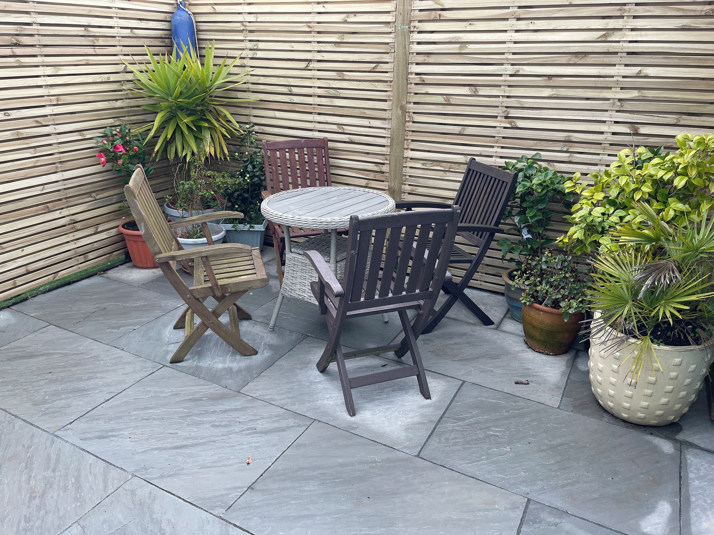 Un patio con una mesa redonda y cuatro sillas, rodeado de macetas con plantas y una cerca de madera.
