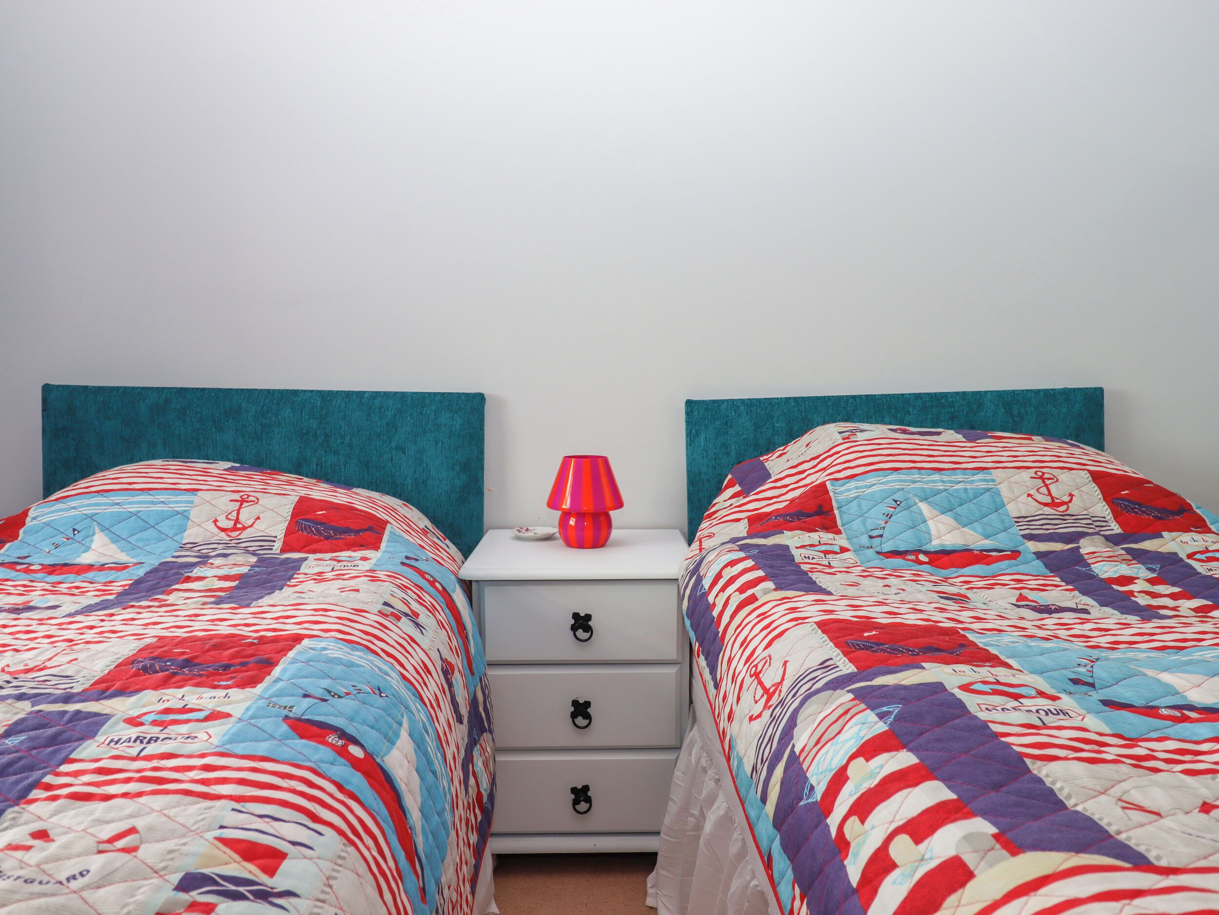Camera da letto con due letti, testiere color turchese, un comodino bianco con cassetti e una lampada rossa.