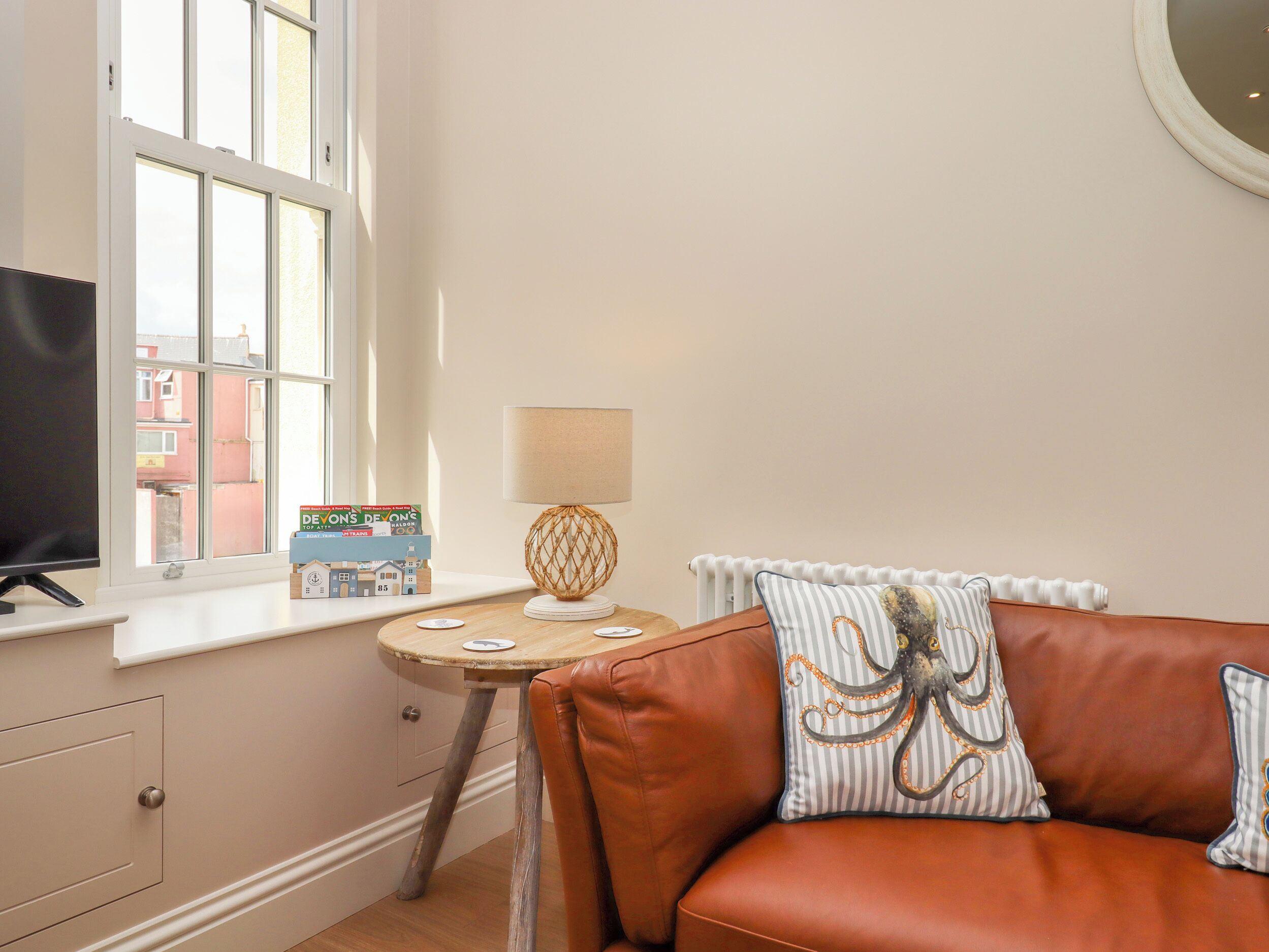A cozy room with a brown leather sofa, a small round table with a lamp, a television on a stand, and a window with a view of buildings outside.