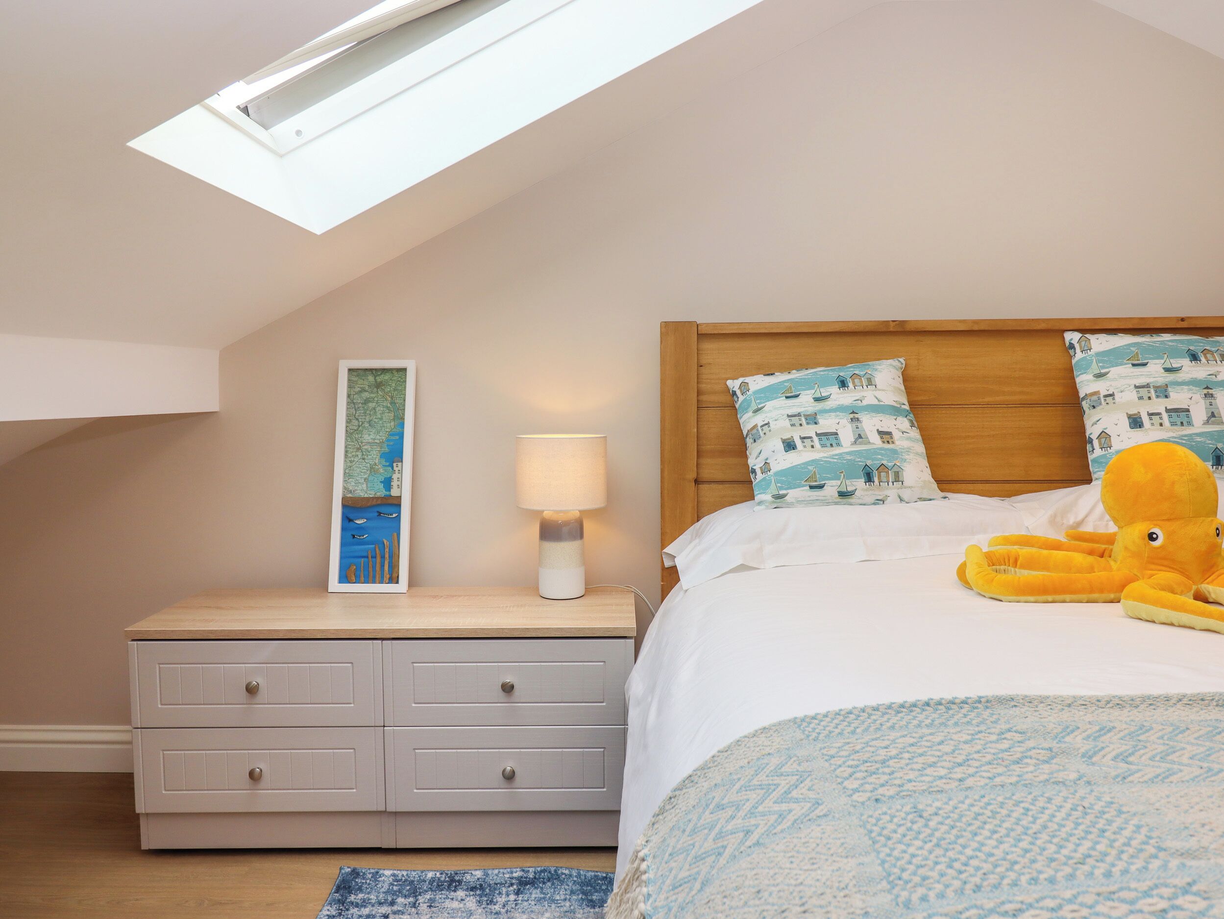 A bedroom with a sloped ceiling, a wooden headboard, a bed with white bedding, a lamp, a map artwork, a plush octopus toy, and a blue patterned throw.