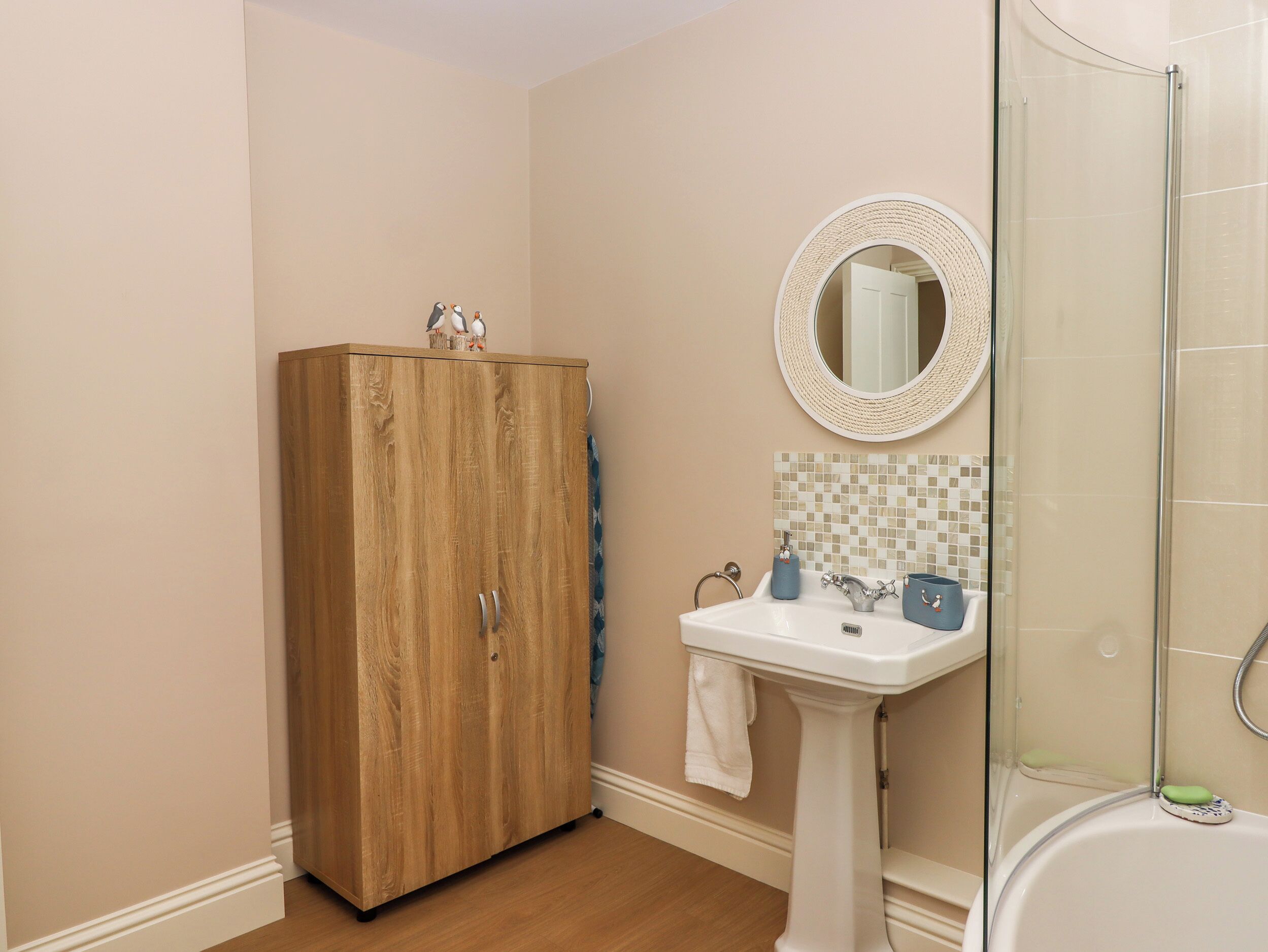 A bathroom with a wooden wardrobe, a white pedestal sink, a round mirror, and a glass shower enclosure.