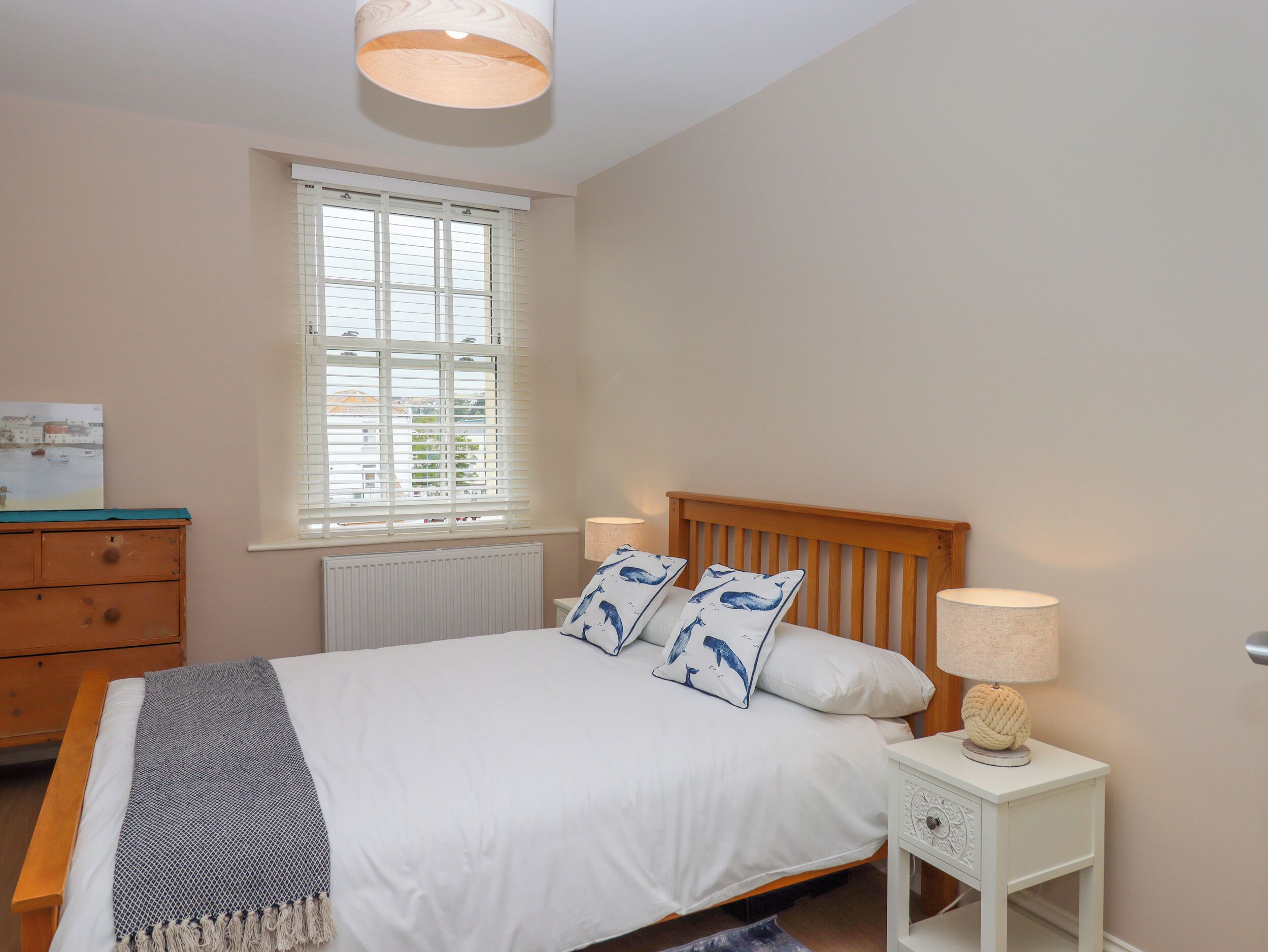 Un dormitorio con una cama de madera, ropa de cama blanca, una mesita de noche de madera con una lámpara, un aparador y una ventana con persianas.