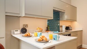 A modern kitchen with white cabinets, a blue backsplash, and a small island with croissants, orange juice, and a coffee maker. - 2 Sea Otter House (Teignmouth)