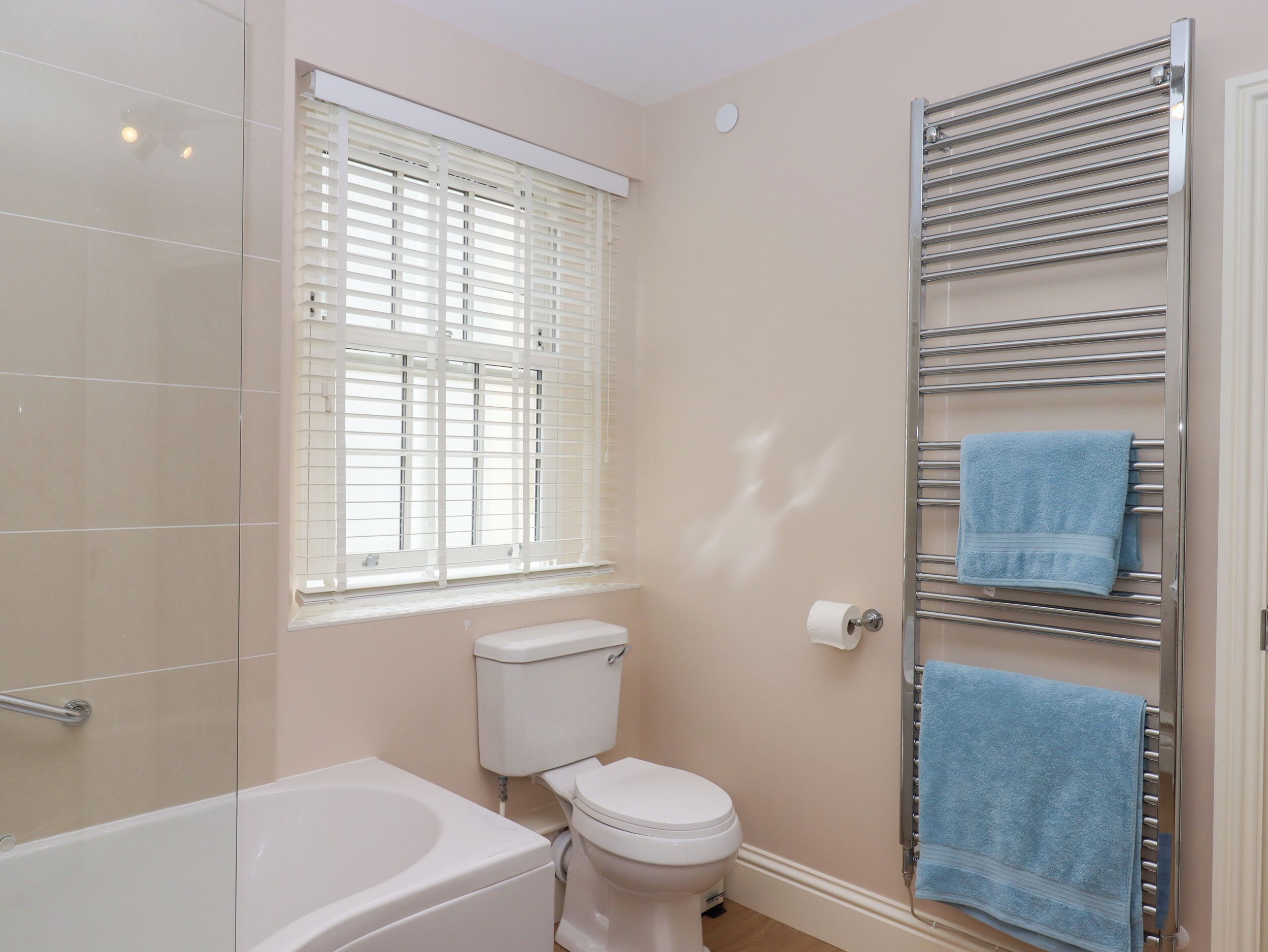 A bathroom with a bathtub, toilet, window with blinds, and a towel rack with towels.