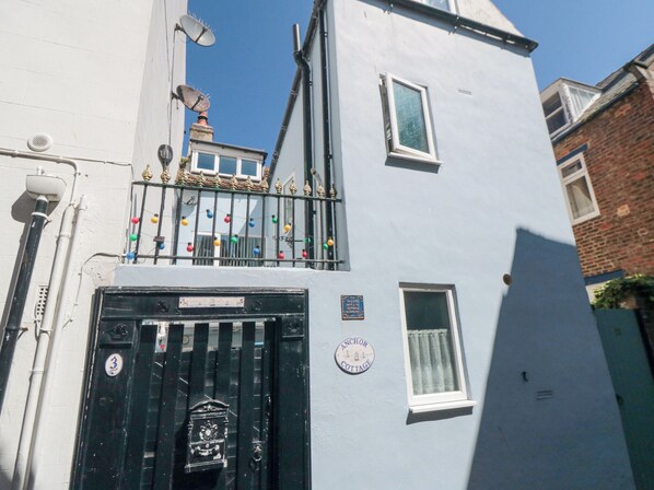 A two-story building with a black gate, a small balcony with colorful lights, and a sign indicating it is a cottage. - 3 Anchor Cottage (Whitby)